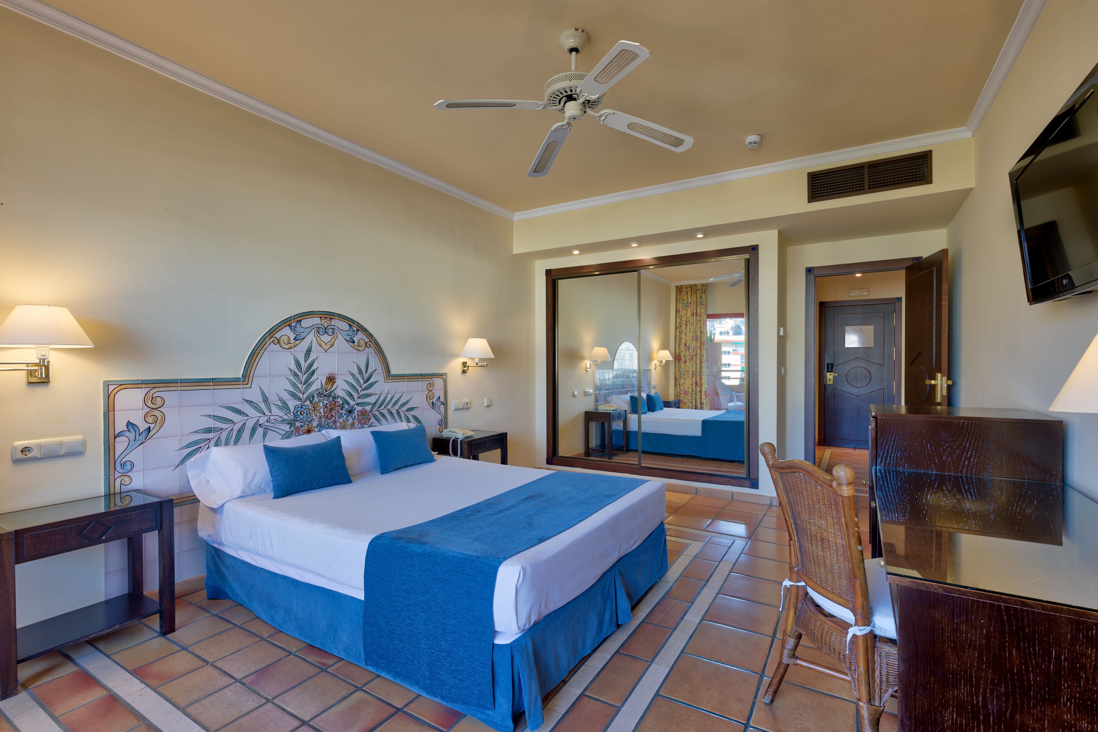 A hotel room with a large bed, a ceiling fan, a TV, and a mirror.