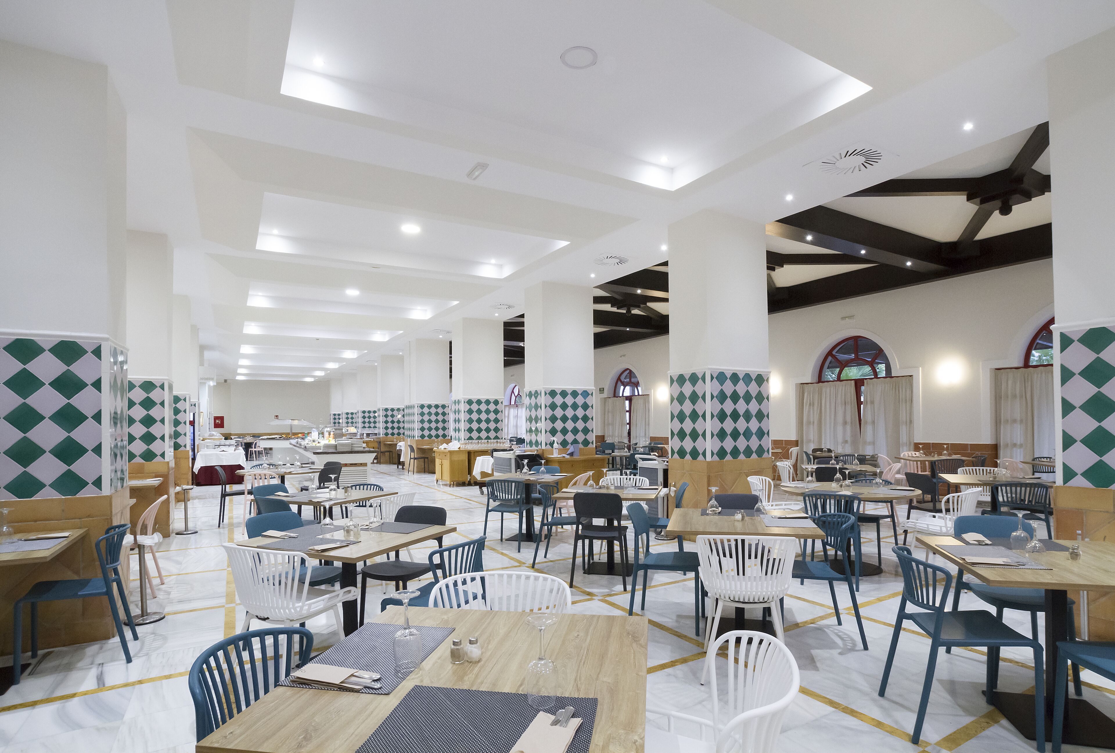 A spacious dining area with multiple tables and chairs, high ceilings with recessed lighting, and patterned wall tiles.