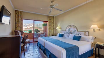 A hotel room with a large bed, a ceiling fan, a desk, and a view of the city through the balcony doors.