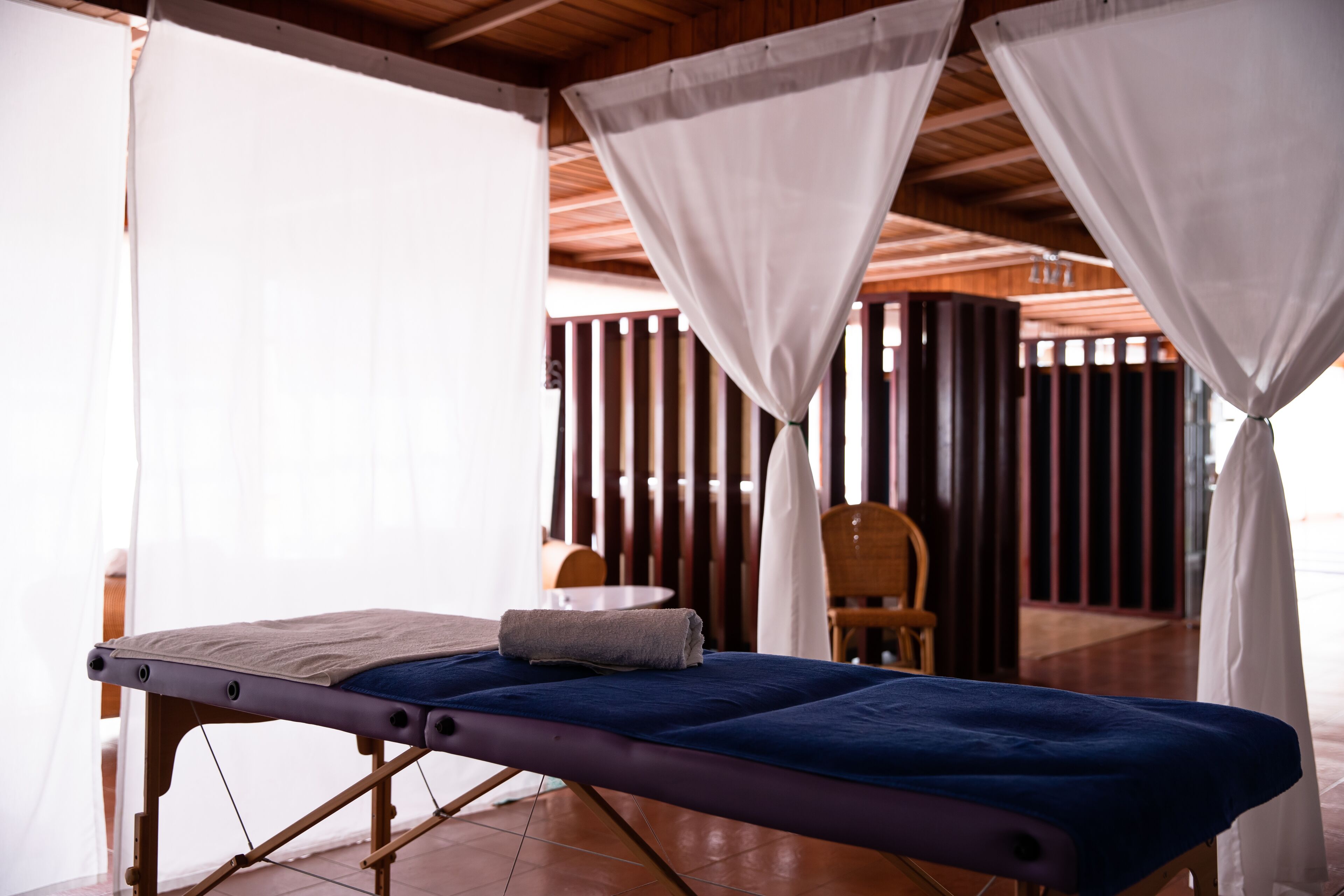 A massage table with a blue mat, a towel, and a wooden frame in a room with wooden beams and white curtains.