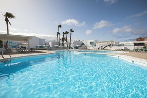 Une piscine d’un bleu éclatant, avec un espace ombragé, des palmiers et un bâtiment blanc doté d’un espace de repos abrité.