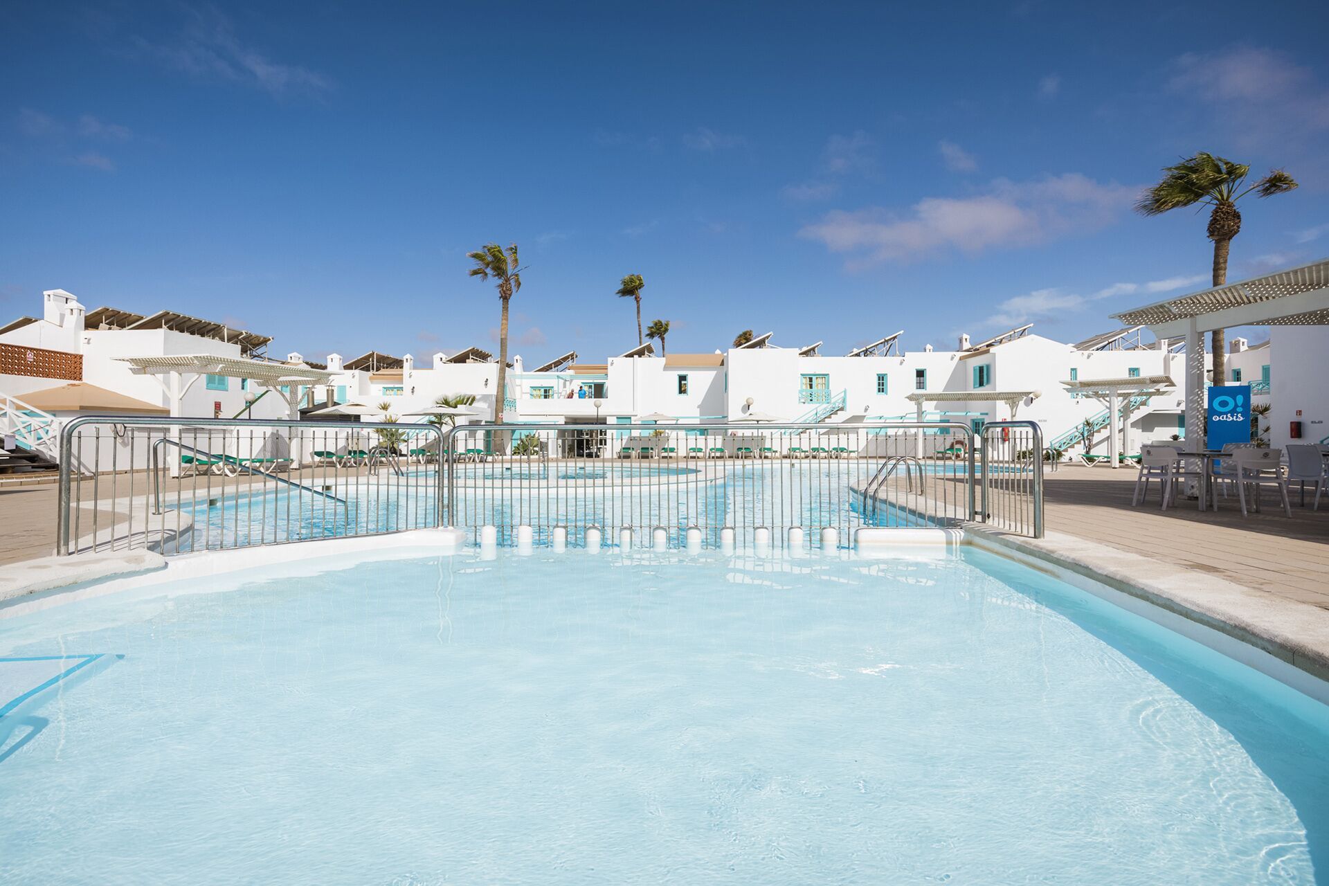 A swimming pool with a metal railing, surrounded by white buildings with blue accents and palm trees.