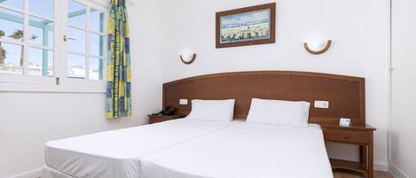 A double bed with white bedding, a wooden headboard, bedside tables with lamps, a painting on the wall, and a window with curtains.