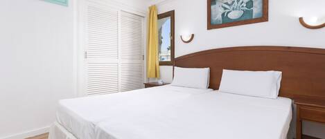 A neatly made bed with white linens, a wooden headboard, and a single bedside table with a lamp.