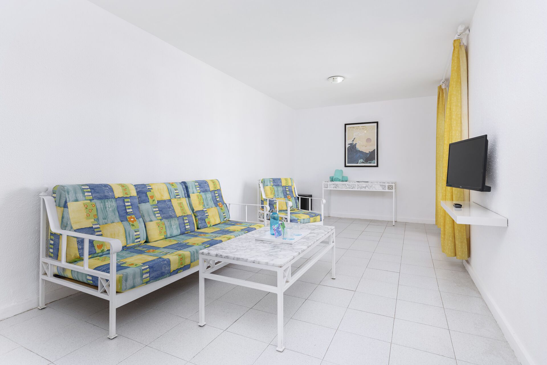 A room with a white sofa, a matching armchair, a small table, a TV on a stand, and a painting on the wall.