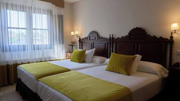 Habitación de hotel con dos camas, cada una con cubrecama verde, ropa de cama blanca y almohadas a juego. Hay dos lámparas de noche y una mesita de noche con lámpara. La habitación tiene un ventanal con cortinas de gasa y vistas al exterior.