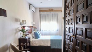 Un dormitorio con cama, mesita de noche con lámpara, una silla y ventana con cortinas traslúcidas.