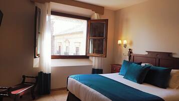 A hotel room with a large bed, a wooden nightstand, a bench, and a window with curtains.