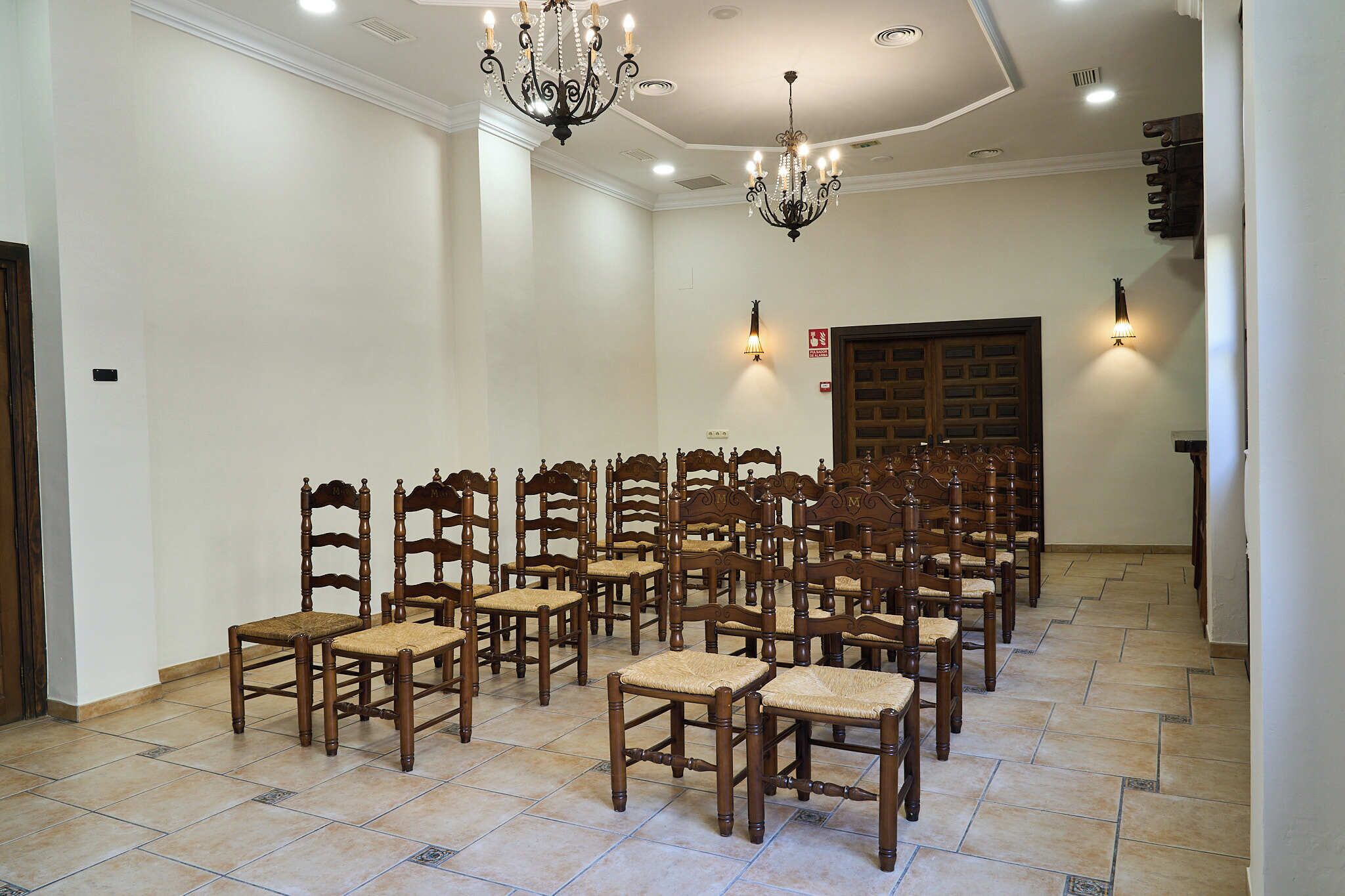 A room with multiple wooden chairs arranged in rows, a chandelier, and wall-mounted lights.