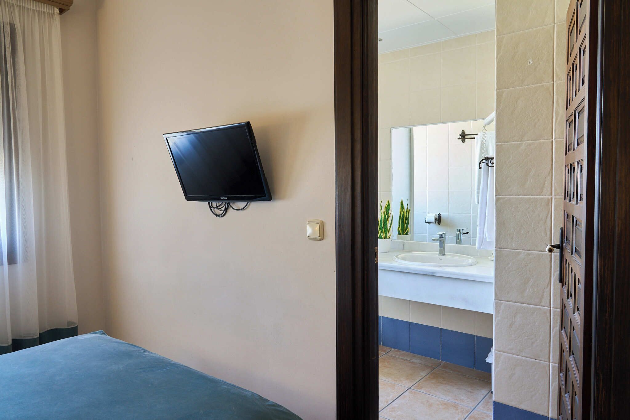 A hotel room with a bed, a mounted television, a bathroom with a sink and mirror, and a door leading to another room.