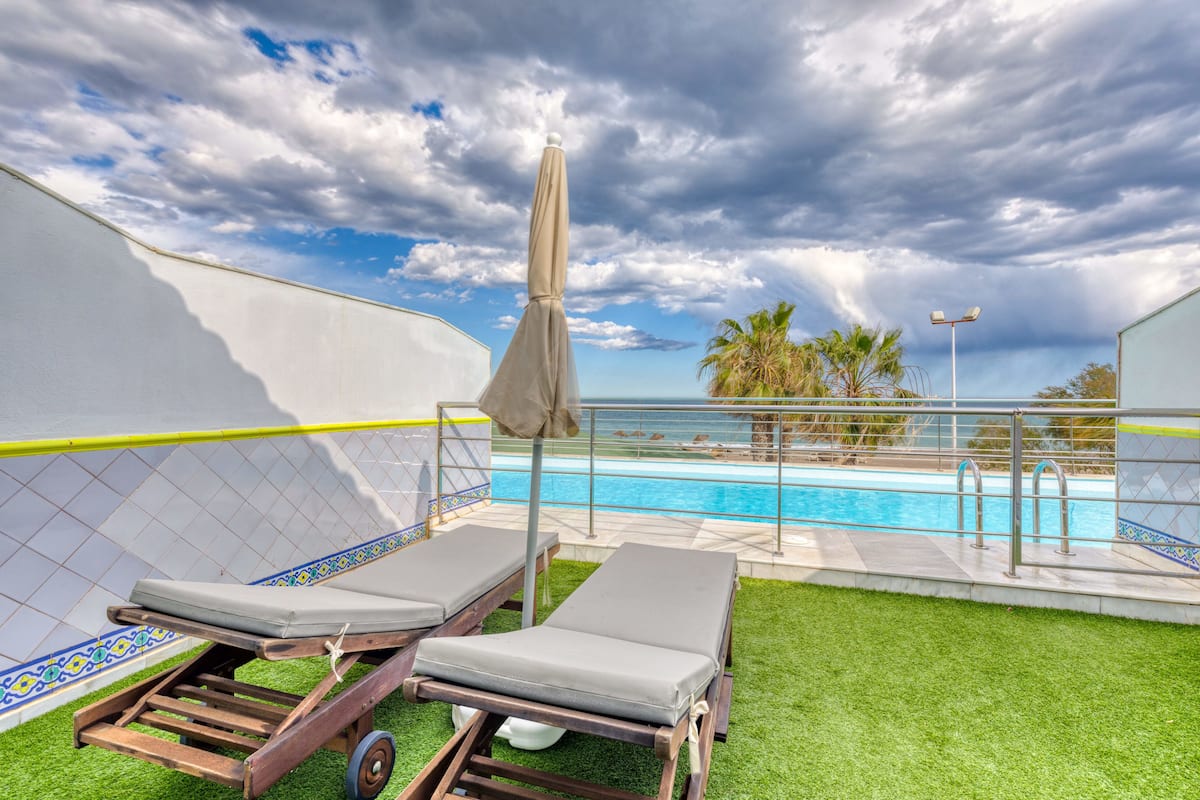A poolside area with two lounge chairs, a covered umbrella, and a view of the pool and palm trees under a cloudy sky.