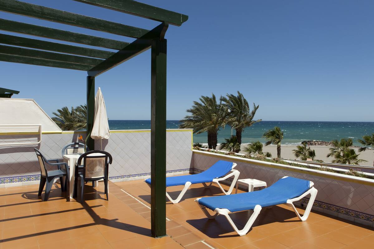 A terrace with two blue lounge chairs, a table with chairs, and a grill.