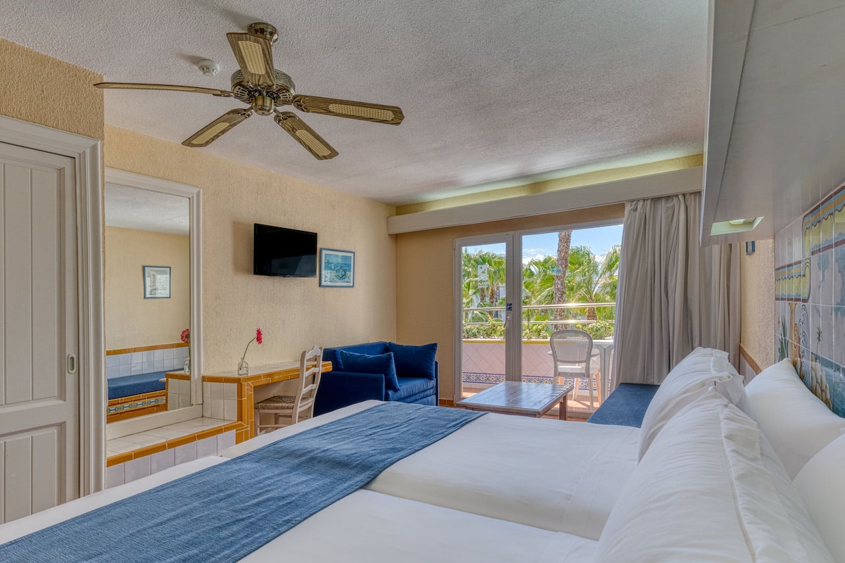 A hotel room with a bed, a ceiling fan, a TV, a balcony with a chair, and a view of palm trees.