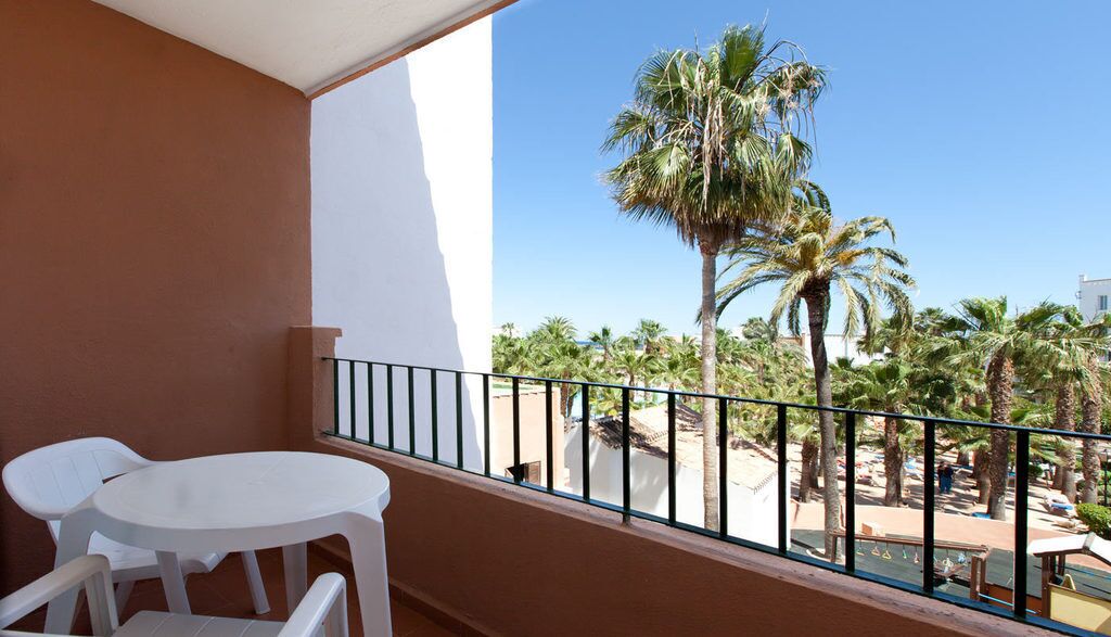 A balcony with a white table and chairs, a metal railing, and a view of palm trees and a beach.