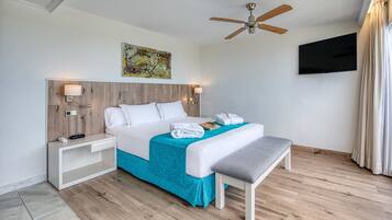 Habitación de hotel con cabecera de madera, televisor de pantalla plana, ventilador de techo y un amplio ventanal con cortinas.