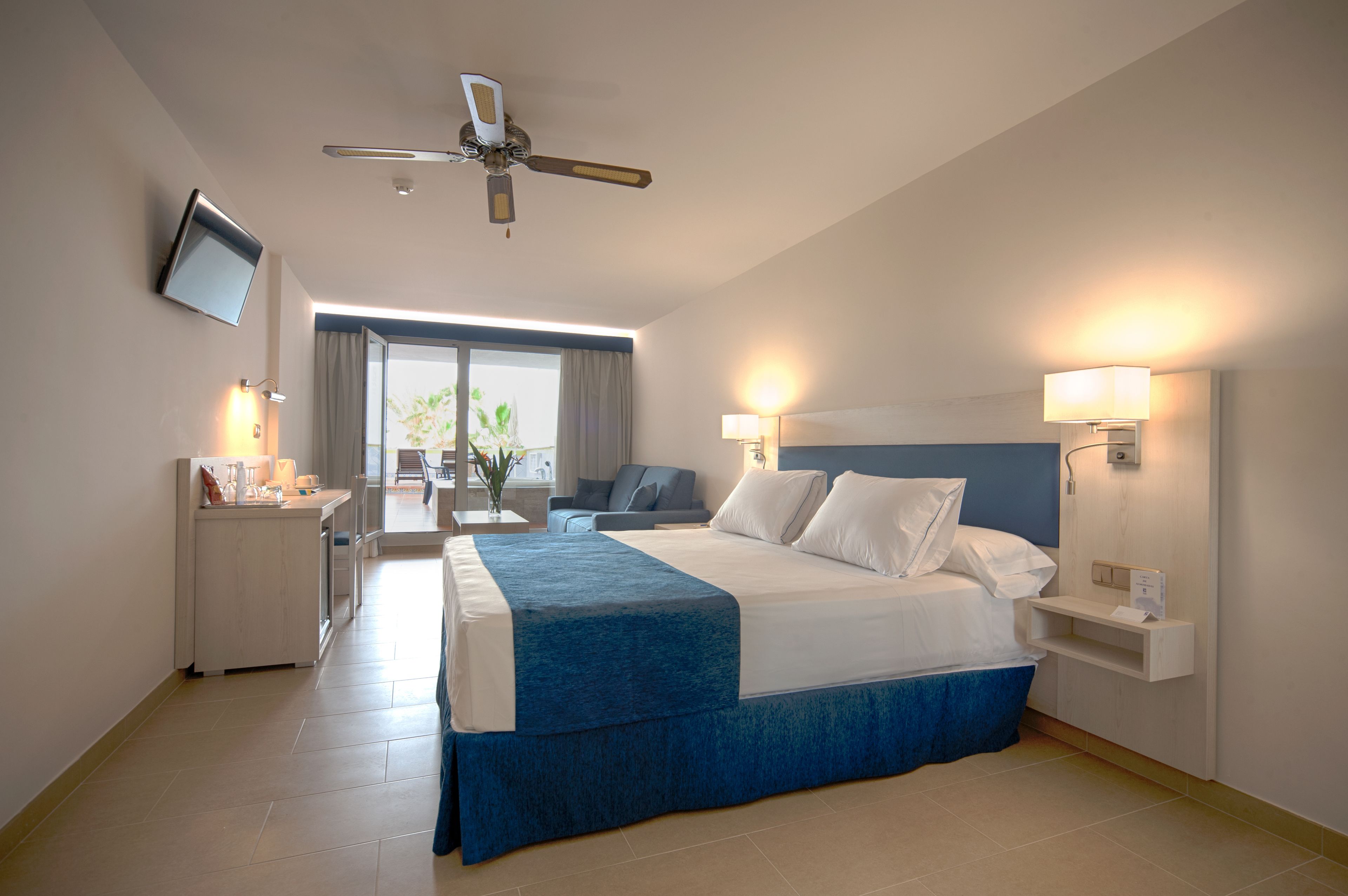 A hotel room with a bed, bedside tables, a TV, a ceiling fan, and a view of the outdoors.