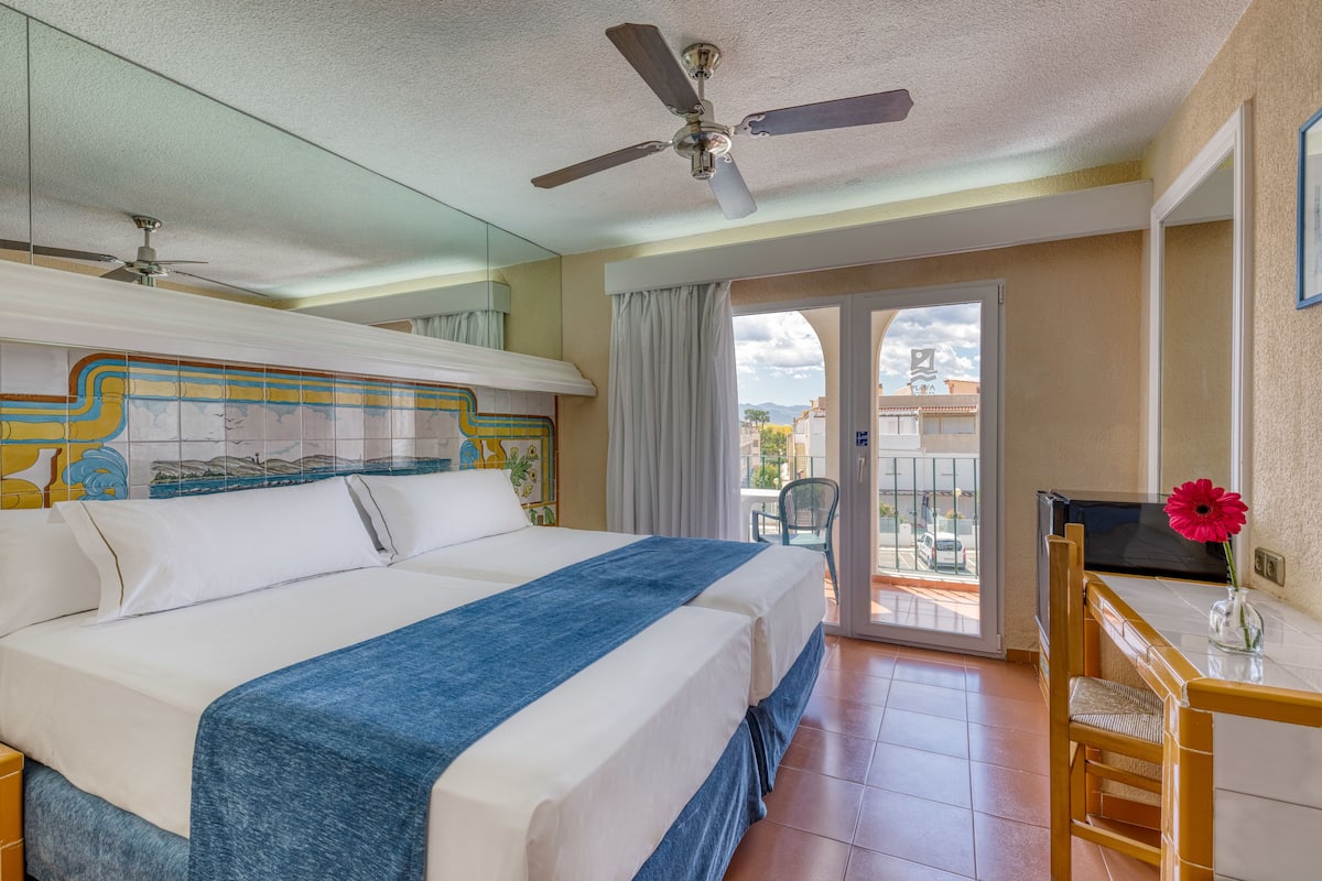 A hotel room with a bed, a desk with a chair, a TV, a ceiling fan, and a view of the outside.
