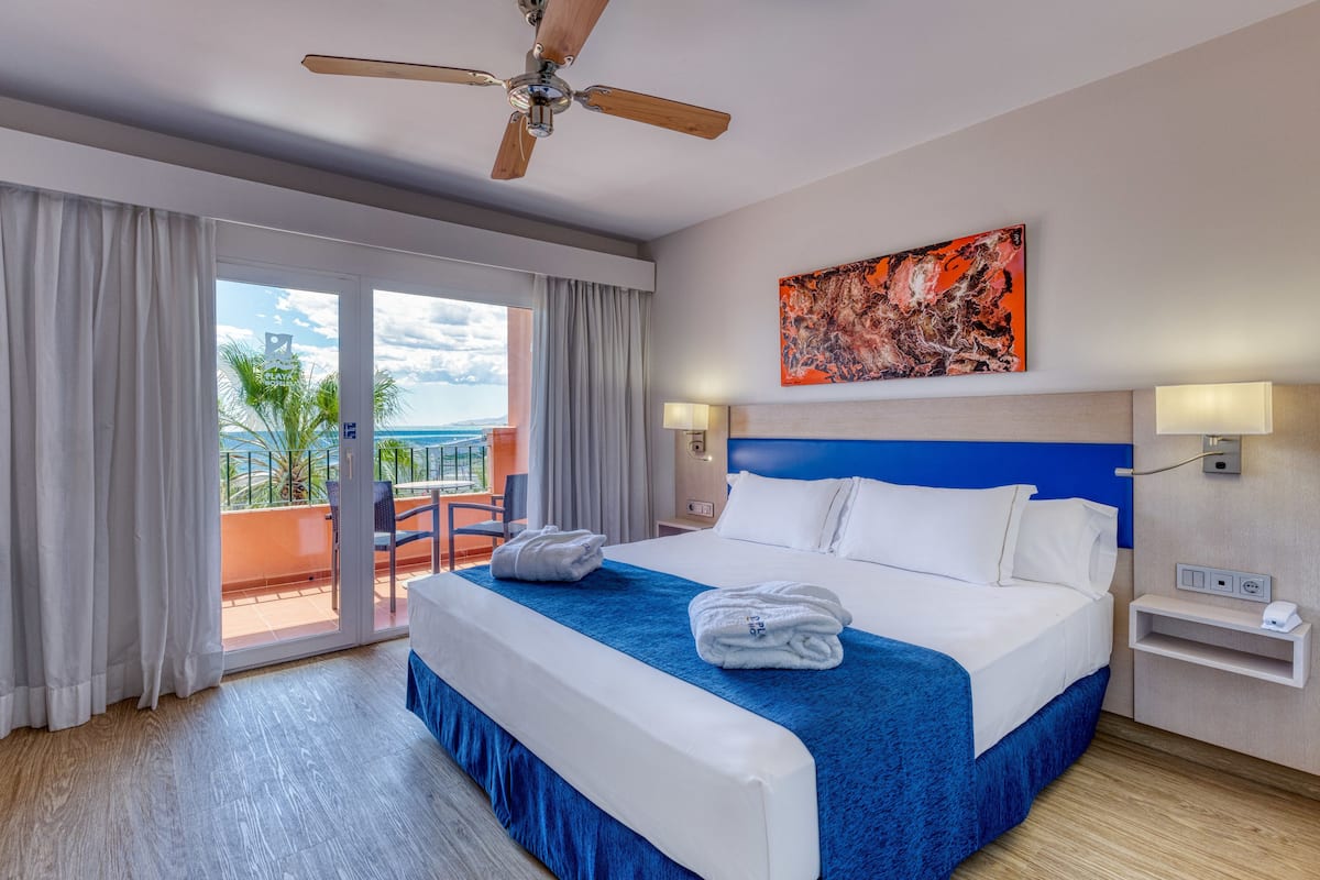 A hotel room with a large bed, a ceiling fan, a balcony with a view of the ocean, and a painting on the wall.