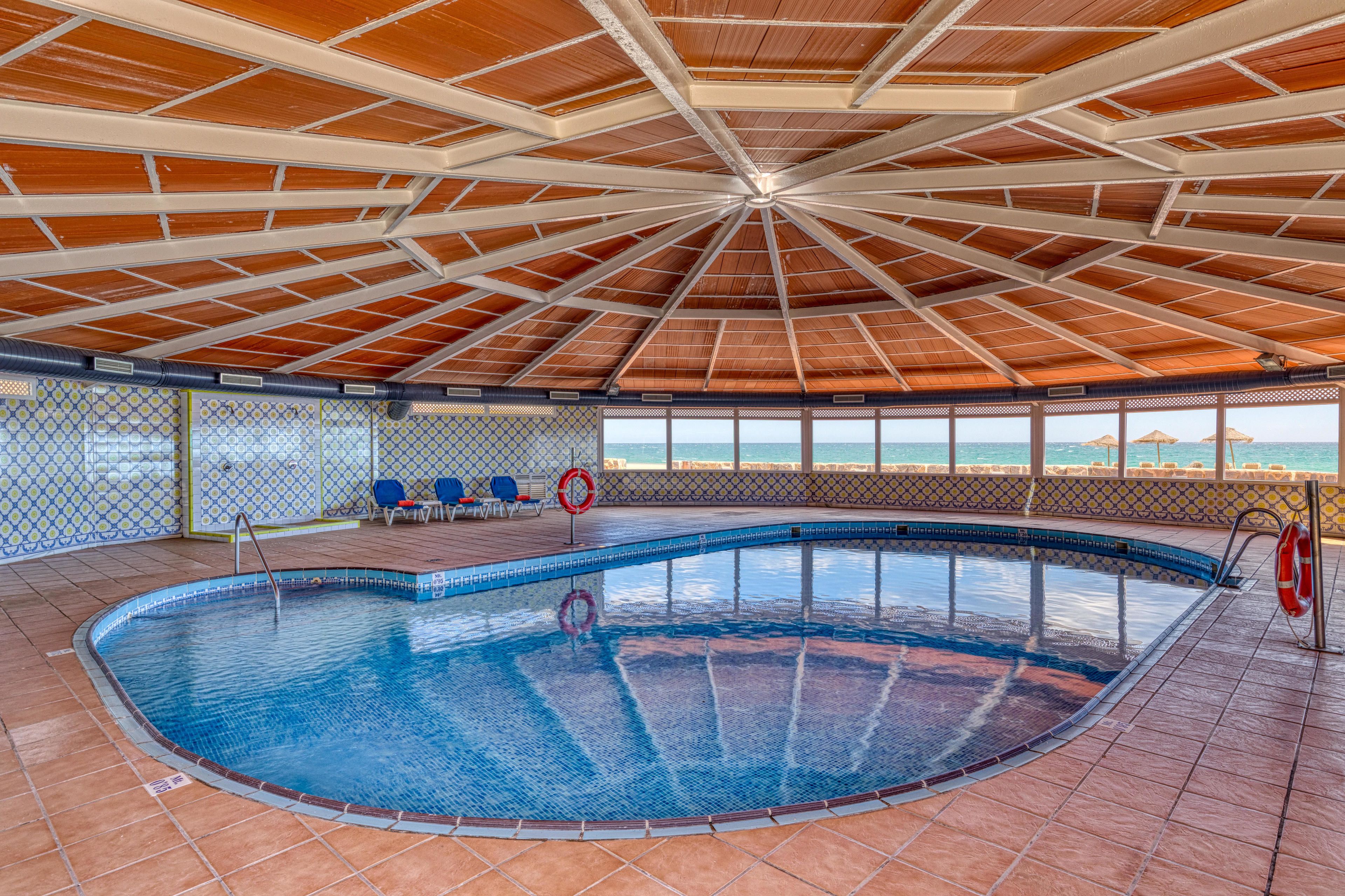 Ein Hallenbad mit Fliesenboden, Holzbalkendecke und Blick auf das Meer durch große Fenster.