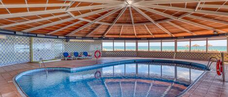 Ein Hallenbad mit Fliesenboden, Holzbalkendecke und Blick auf das Meer durch große Fenster.