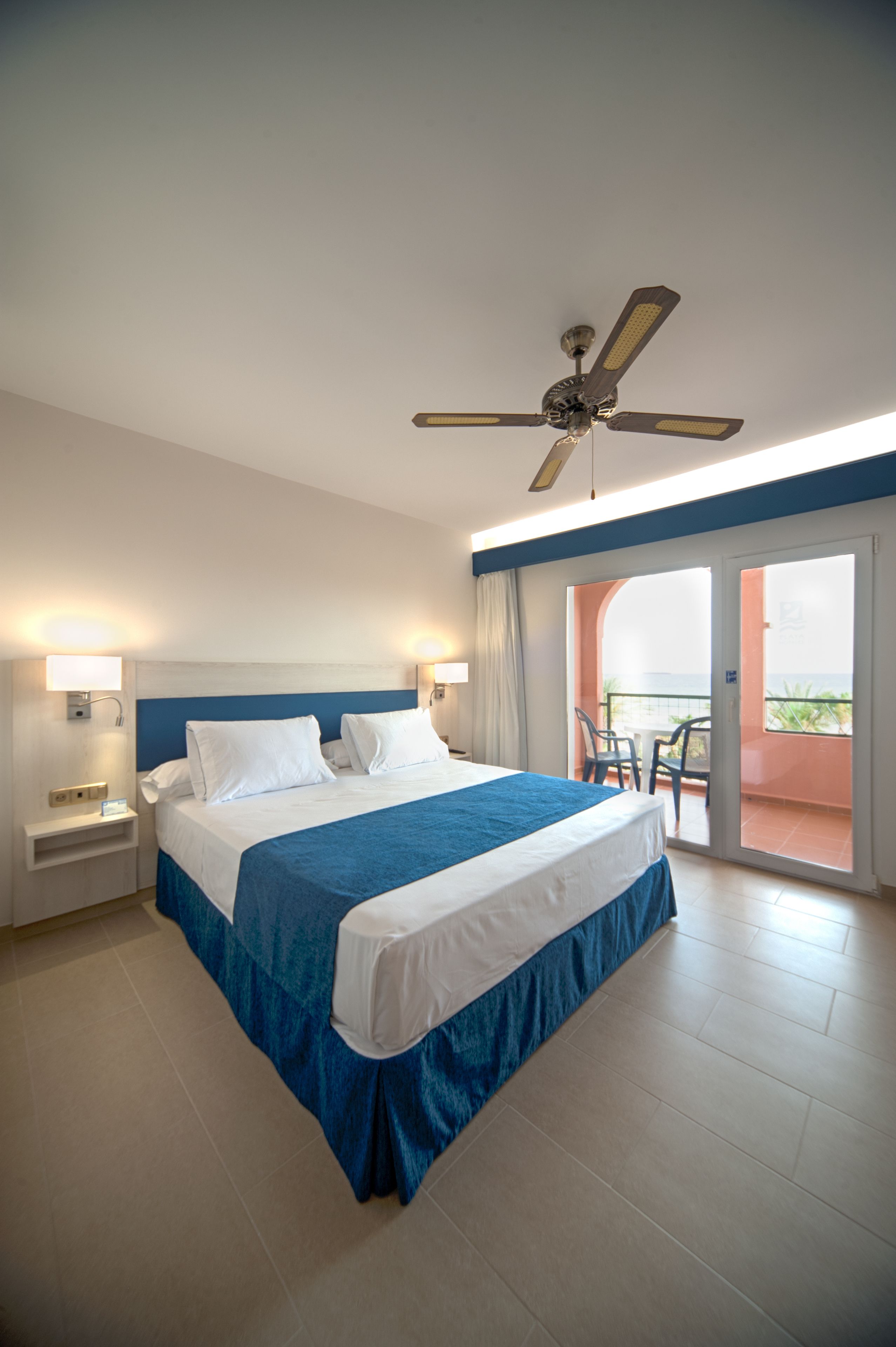 Habitación de hotel con cama, ventilador de techo, mesita de noche, mesa con sillas y balcón con vistas.