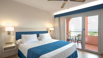 Habitación de hotel con cama, ventilador de techo, mesita de noche, mesa con sillas y balcón con vistas.