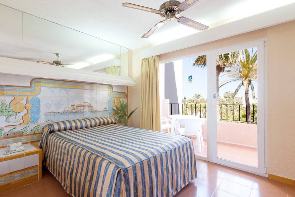 A hotel room with a bed, a ceiling fan, a balcony with chairs, and a view of palm trees.