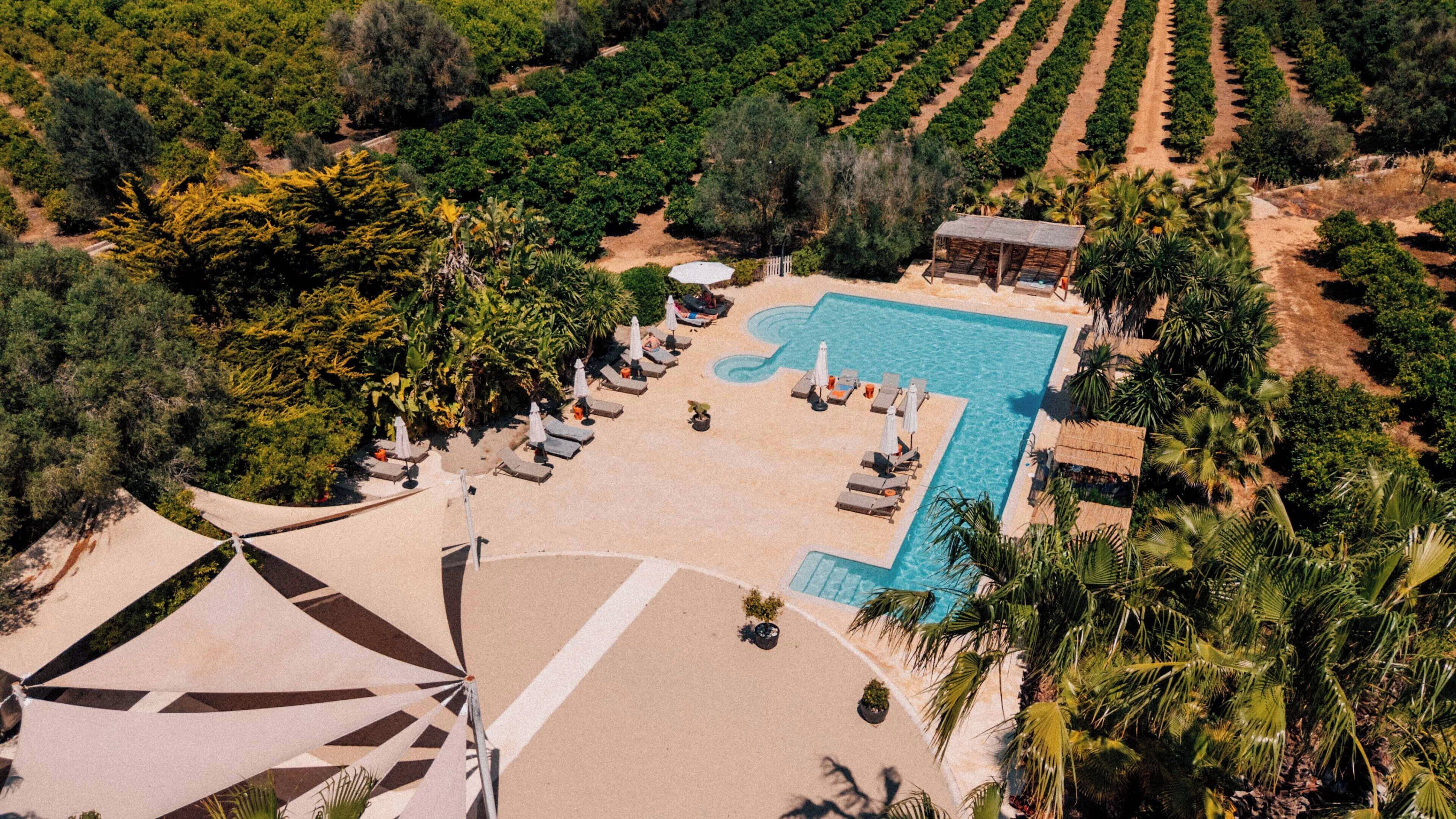 Vue aérienne d’un complexe hôtelier doté d’une piscine, de chaises longues et d’un jardin environnant.
