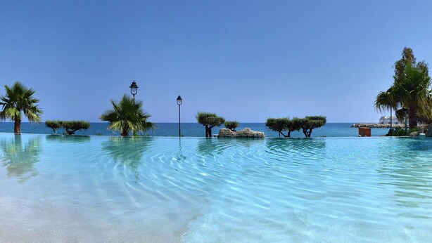 A clear pool with a thatched-roof structure, palm trees, and a rocky outcrop. - GRAN HOTEL LAS FUENTES (Alcala de Xivert)
