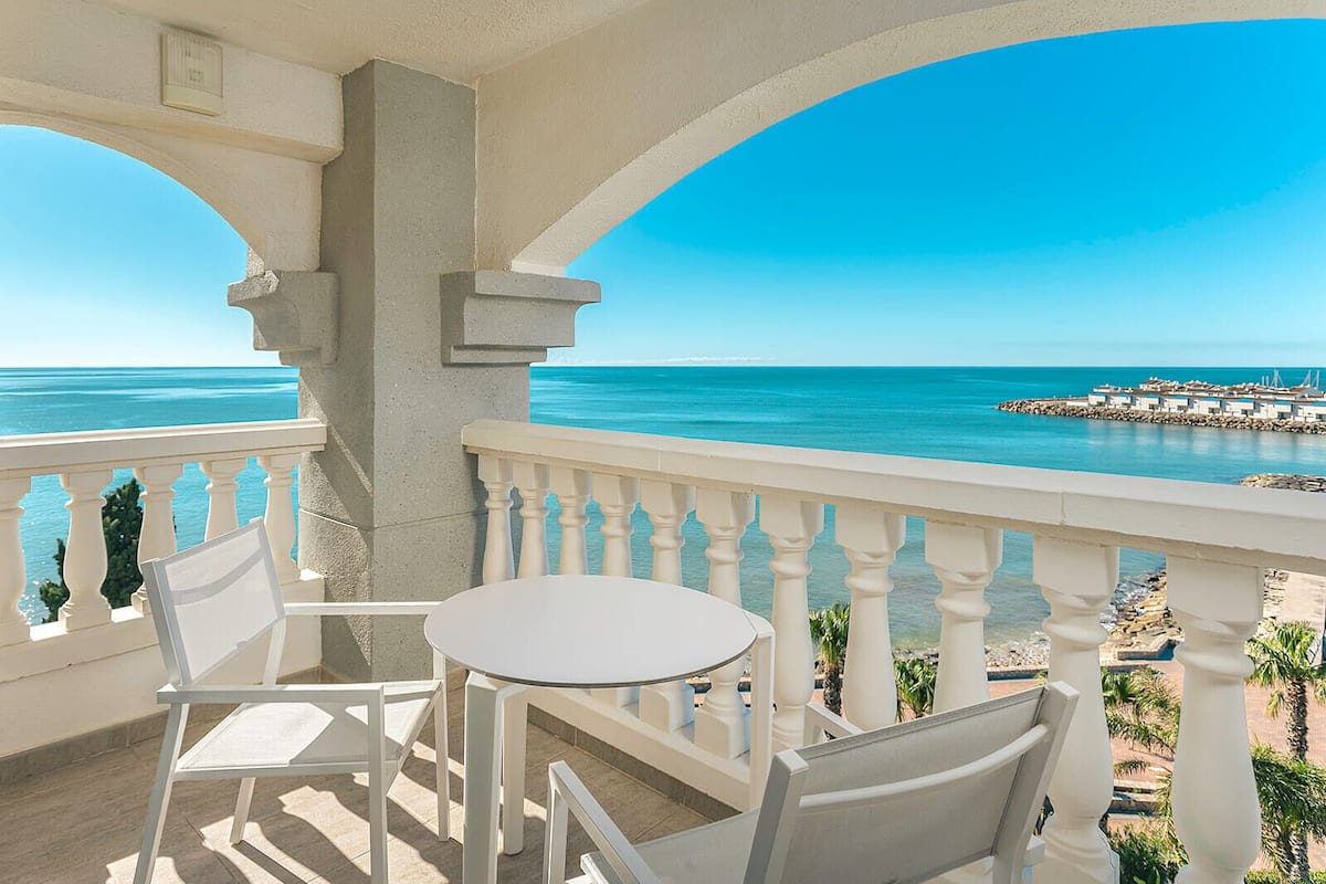 A balcony with a table and chairs, overlooking a beach and a rocky breakwater.