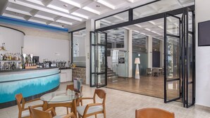 A modern bar area with a glass table and wooden chairs, a counter with various bottles, and a view into a dining area with a staircase. - GRAN HOTEL LAS FUENTES (Alcala de Xivert)