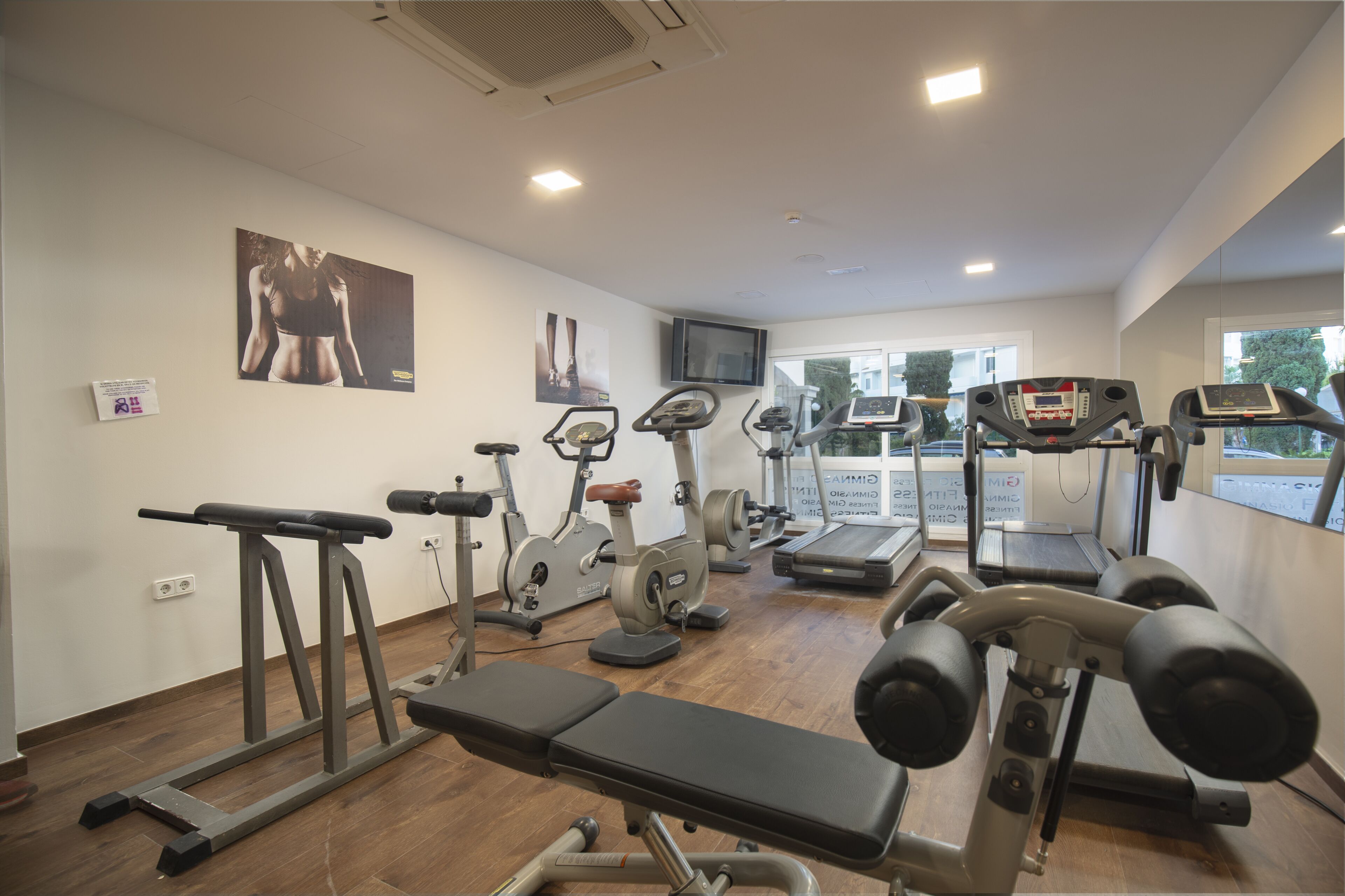 A gym with various exercise equipment, a television on the wall, and a window with a view of the outside.