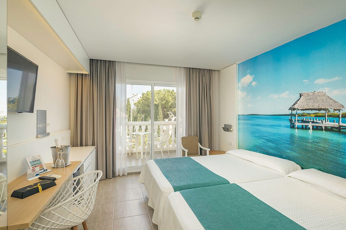 Une chambre d’hôtel avec un lit, un bureau avec un ordinateur, une chaise, une télévision et un balcon donnant sur la mer.