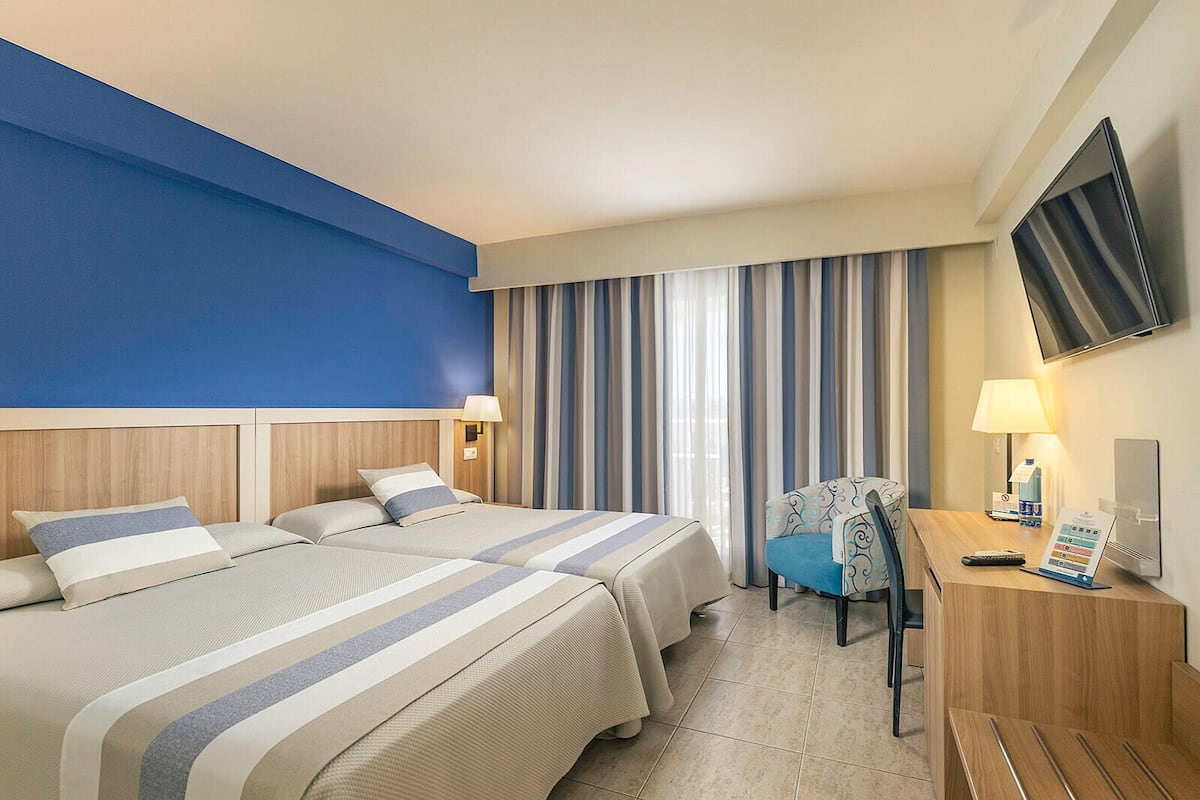 Une chambre d’hôtel avec deux lits, un bureau, une chaise, une télévision et une fenêtre avec des rideaux.