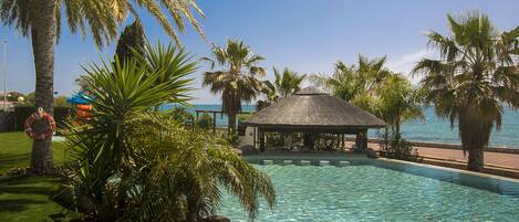 A pool area with a thatched-roof structure, surrounded by palm trees and a clear view of the sea.