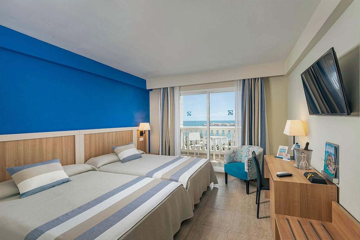 Une chambre d’hôtel avec deux lits, un bureau avec une lampe, une télévision et une vue sur la mer.