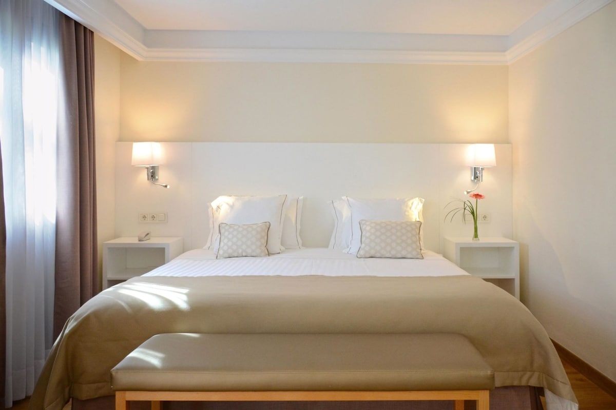 Habitación de hotel con una cama grande, dos mesitas de noche, un banco y una lámpara de pared.