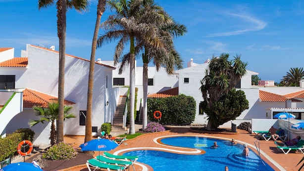 A swimming pool with lounge chairs and umbrellas, surrounded by palm trees and white buildings. - Puerto Caleta (Antigua)