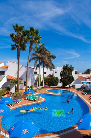 A swimming pool with lounge chairs and umbrellas, surrounded by palm trees and white buildings. - Puerto Caleta (Antigua)