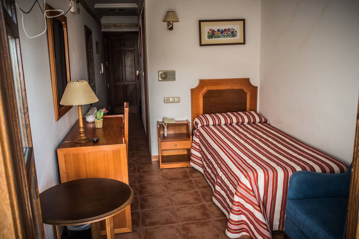 Ein kleines, ordentlich eingerichtetes Schlafzimmer mit einem Einzelbett, einem Nachttisch, einer Lampe und einem Sessel.