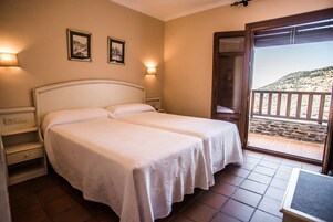 Una cama doble con cubrecama blanco, una mesita de noche y vistas a un balcón con pared de piedra y una ladera.