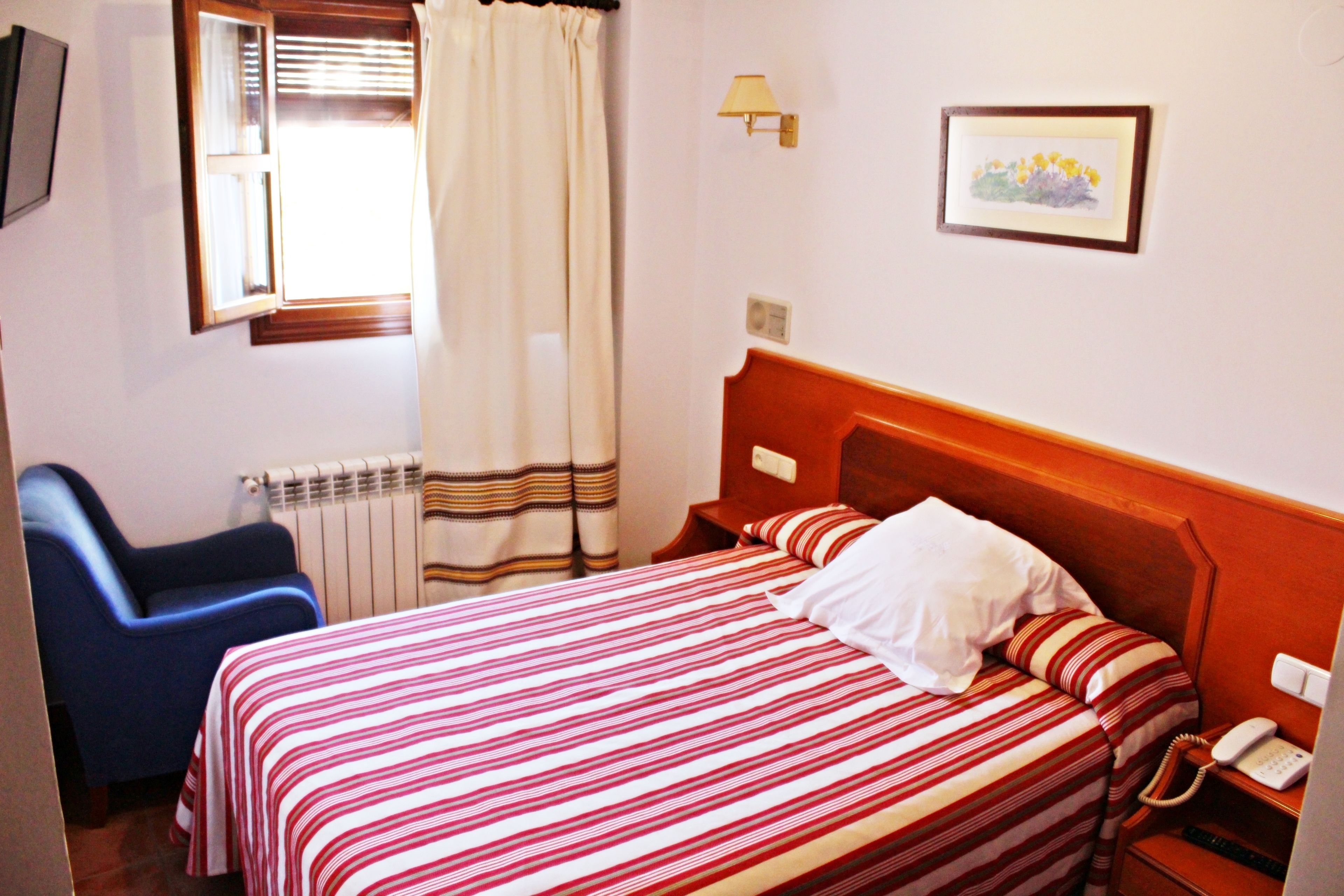 Habitación de hotel con cama, una silla, mesita de noche con teléfono, lámpara y ventana con cortinas.