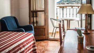 Una habitación de hotel con una cama, un sillón azul, una mesa de comedor con sillas, un balcón con vistas a las colinas y una lámpara sobre la mesa.