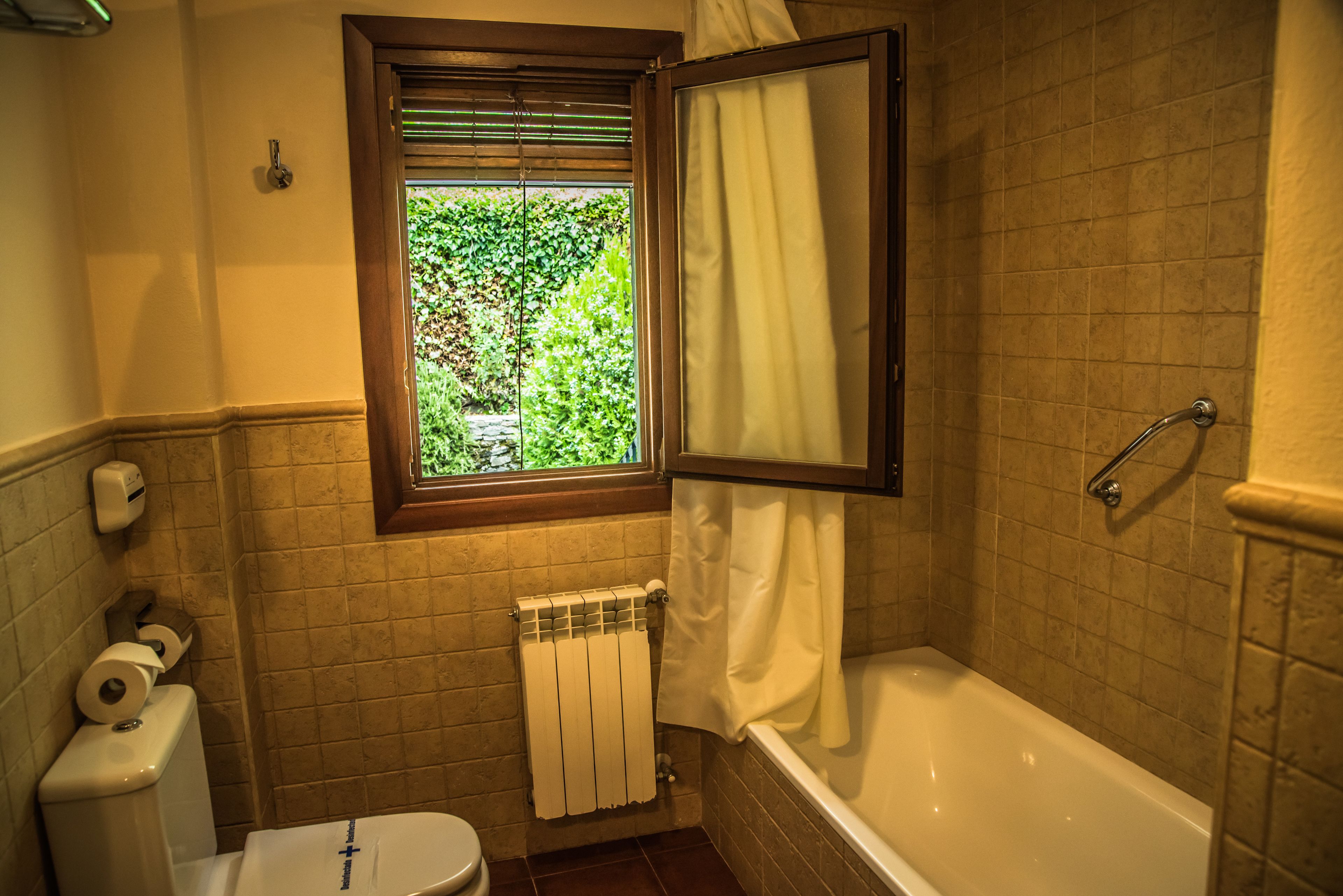 A bathroom with a toilet, a radiator, a window with a view of greenery, and a bathtub.