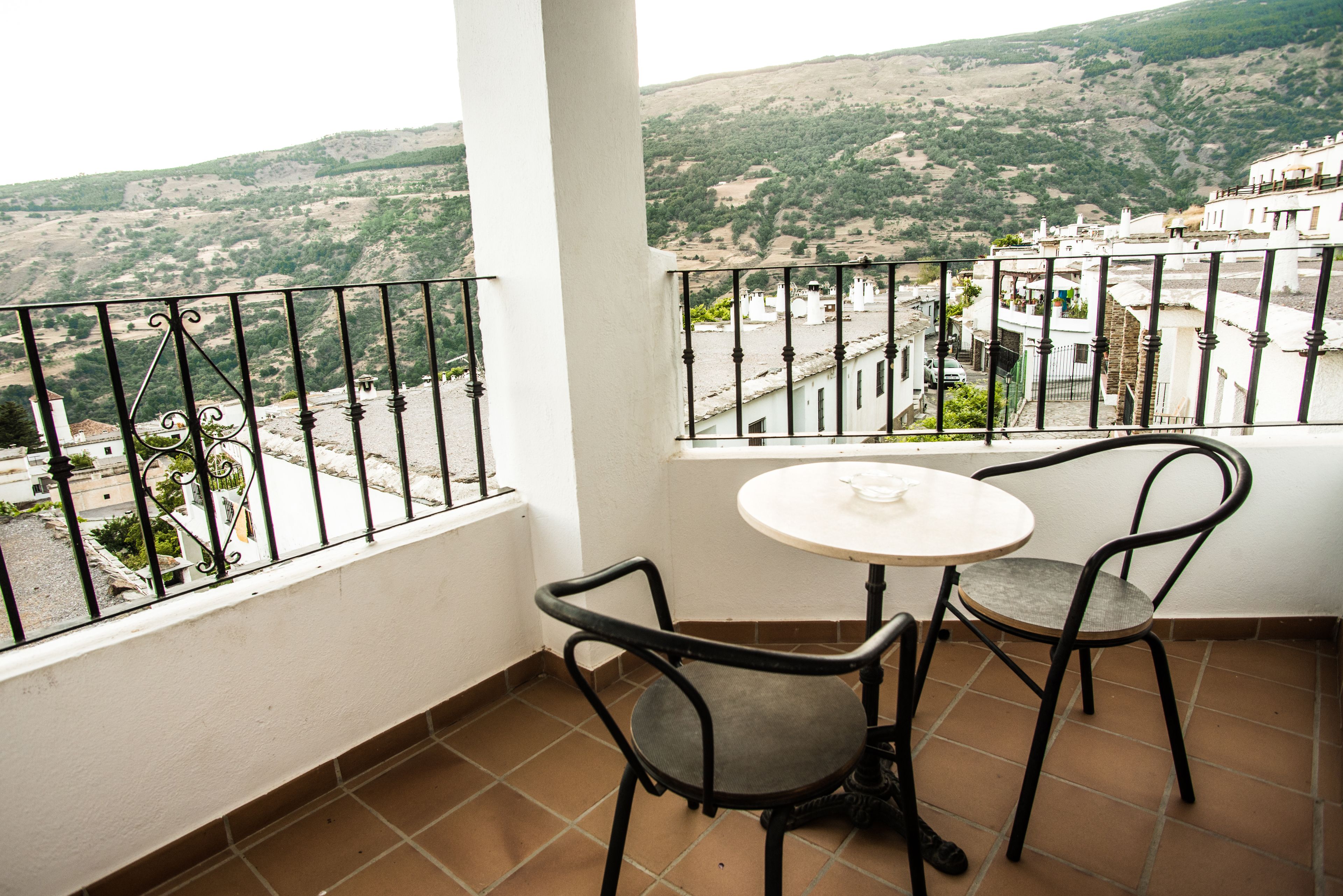 A balcony with a view of a hilly landscape, a small round table, and two chairs.