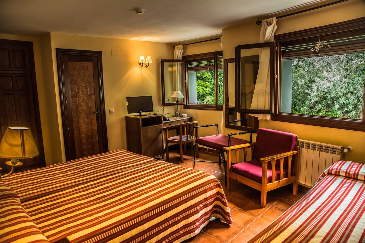 Ein Hotelzimmer mit zwei Betten, einem Schreibtisch mit Computer, einem Stuhl und einem Fenster mit Blick auf Grünflächen.