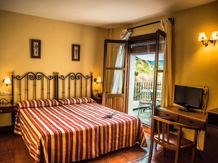 A hotel room with a bed, a desk with a television, and a balcony with a view of the countryside.