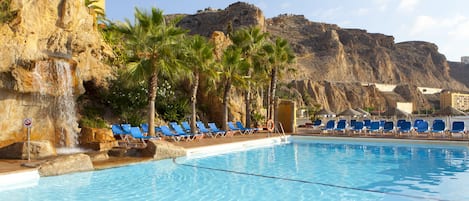 A pool with blue chairs and palm trees, a waterfall, and a rocky cliff in the background.