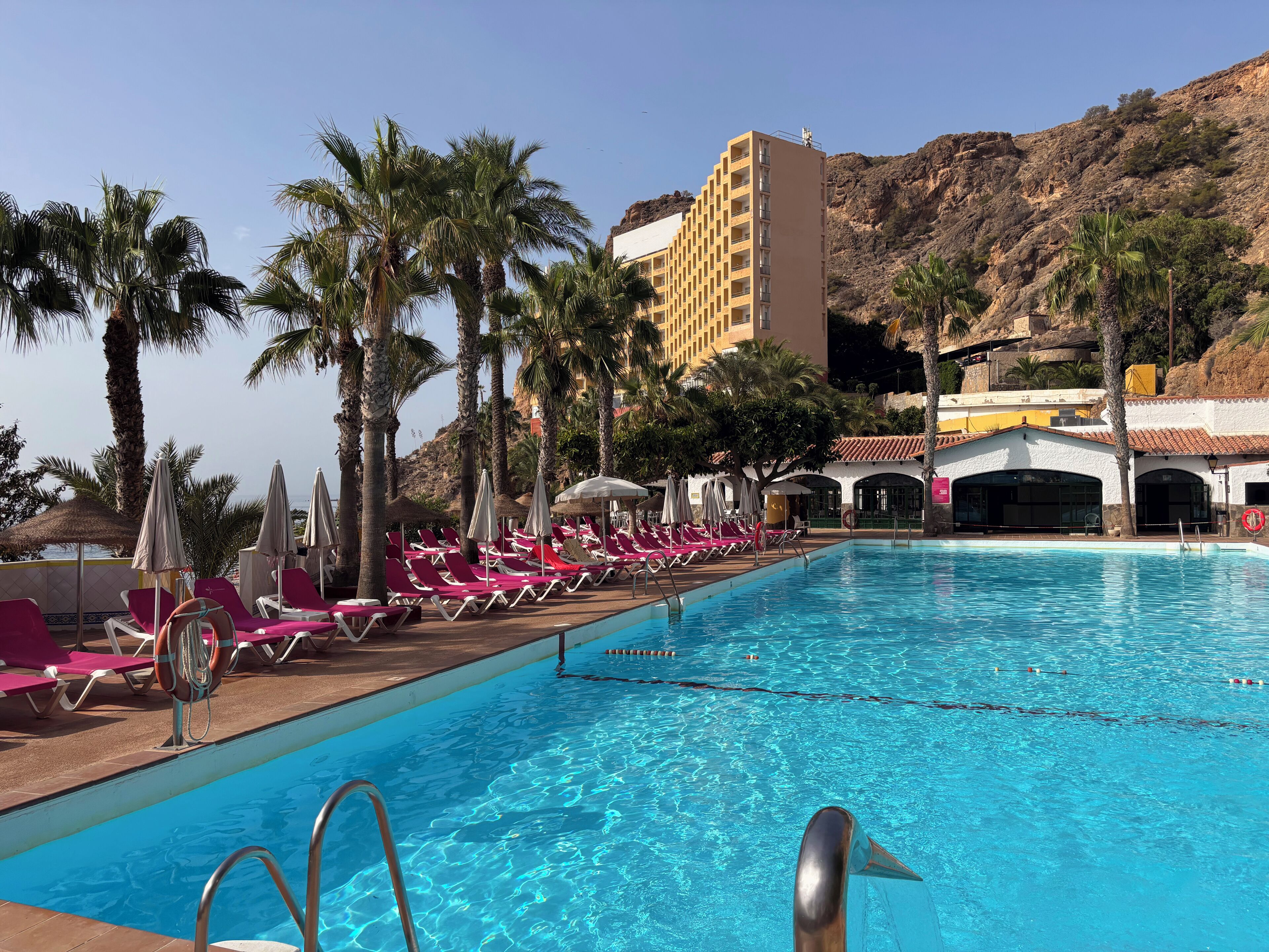 A swimming pool with sun loungers and palm trees, surrounded by buildings and a mountainous backdrop.