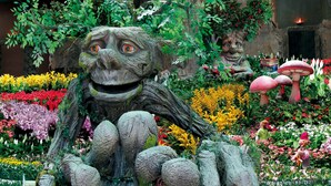 Un jardín con una gran escultura de piedra de un personaje rodeado de diversas flores y plantas.